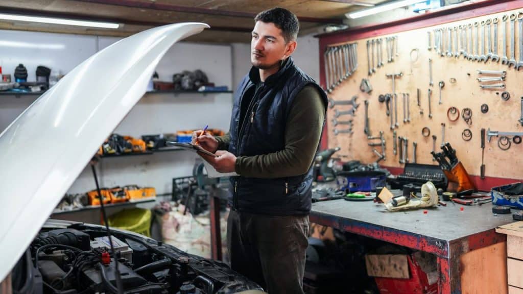 A male mechanic with a clipboard, looking thoughtfully at an open car hood.