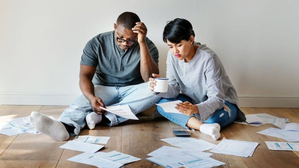 A concerned couple sitting on the floor, surrounded by bills and paperwork.