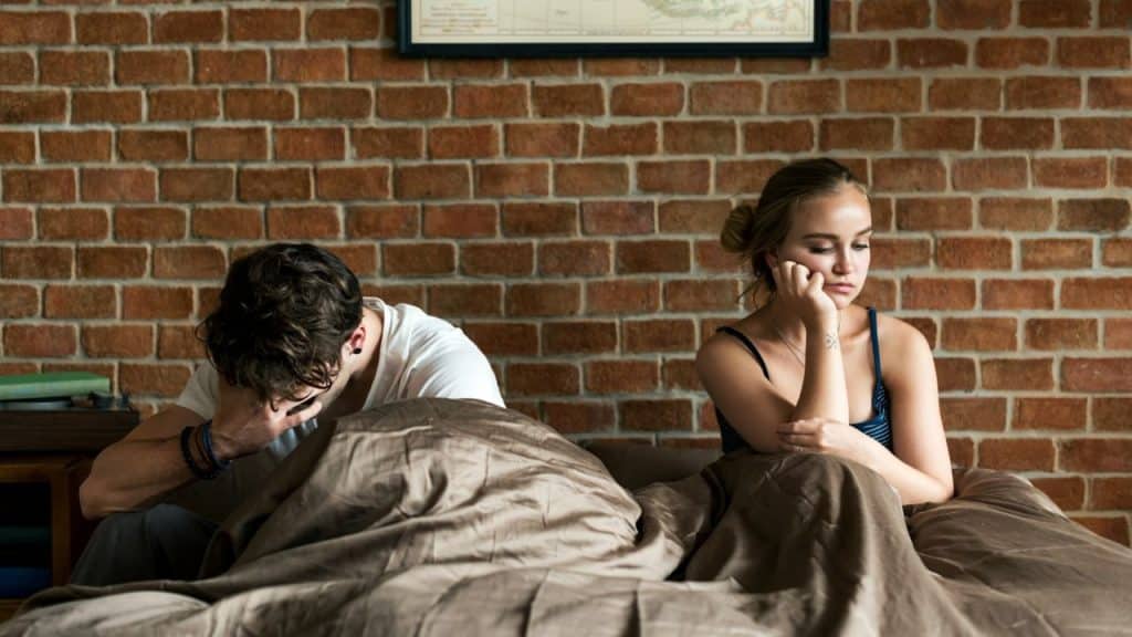 A distressed couple sitting separately on a bed with a brick wall behind them.