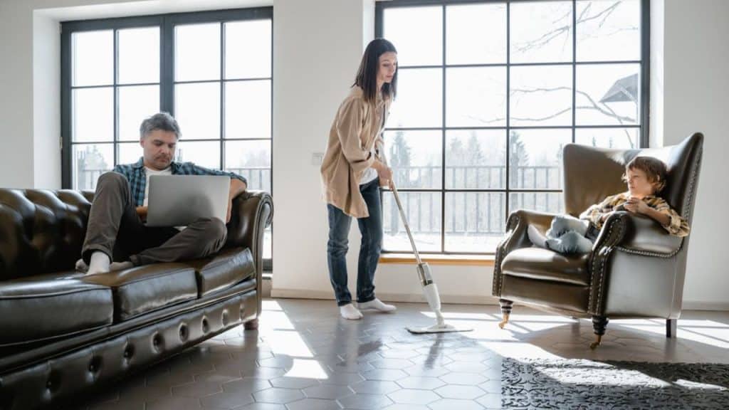 Woman vacuuming while partner sits on phone in the background.