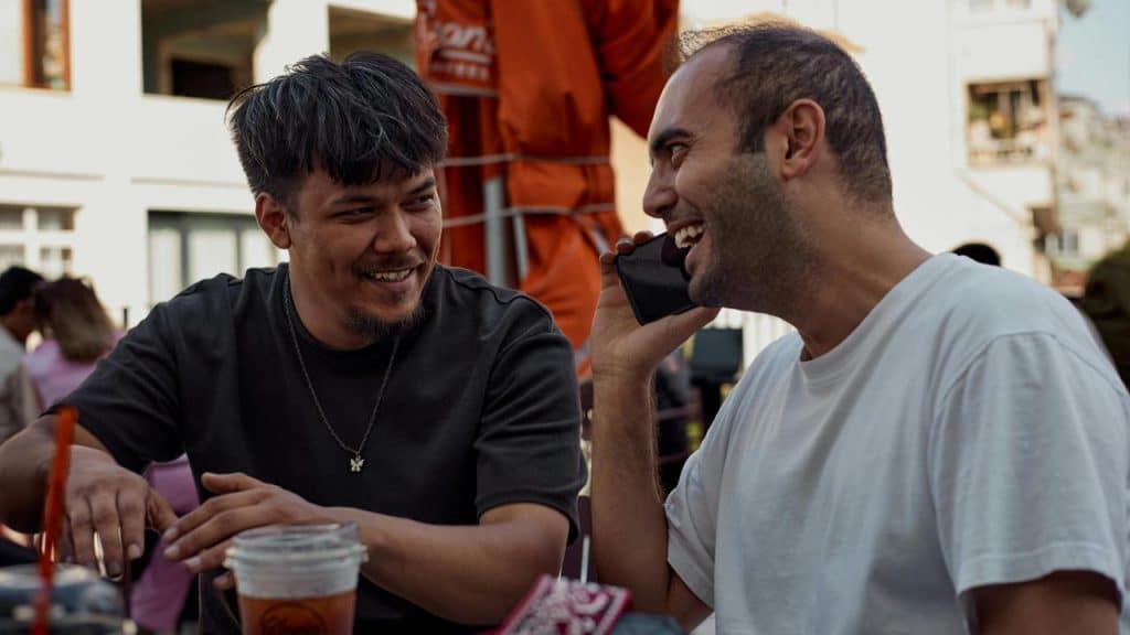 A pair of men laughing at an outdoor café, one talking on a phone.