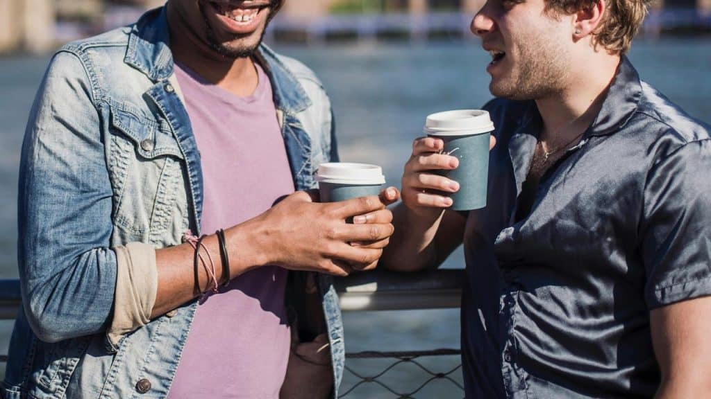A pair of men holding coffee cups and chatting by a riverside railing.