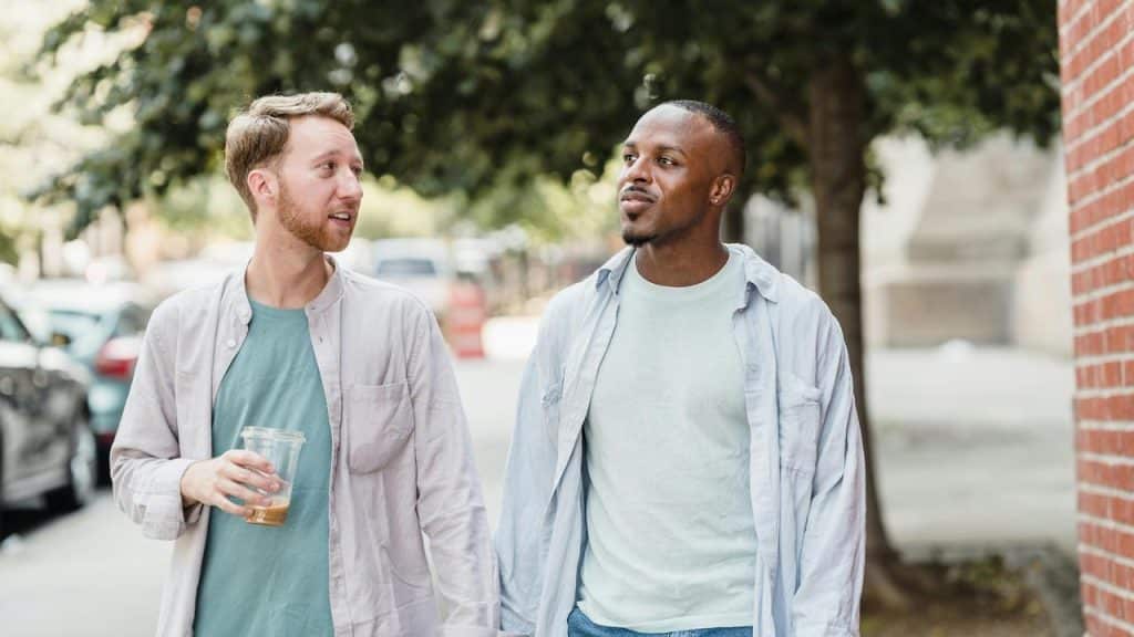A pair of men walking on a sidewalk, one holding an iced coffee.