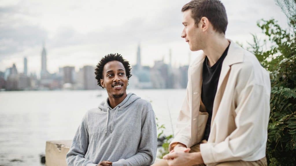 A pair of men talking by a river with a city skyline behind.