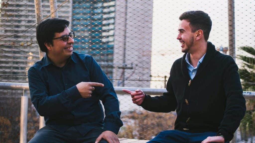 A pair of men sitting outdoors, pointing at each other and laughing.