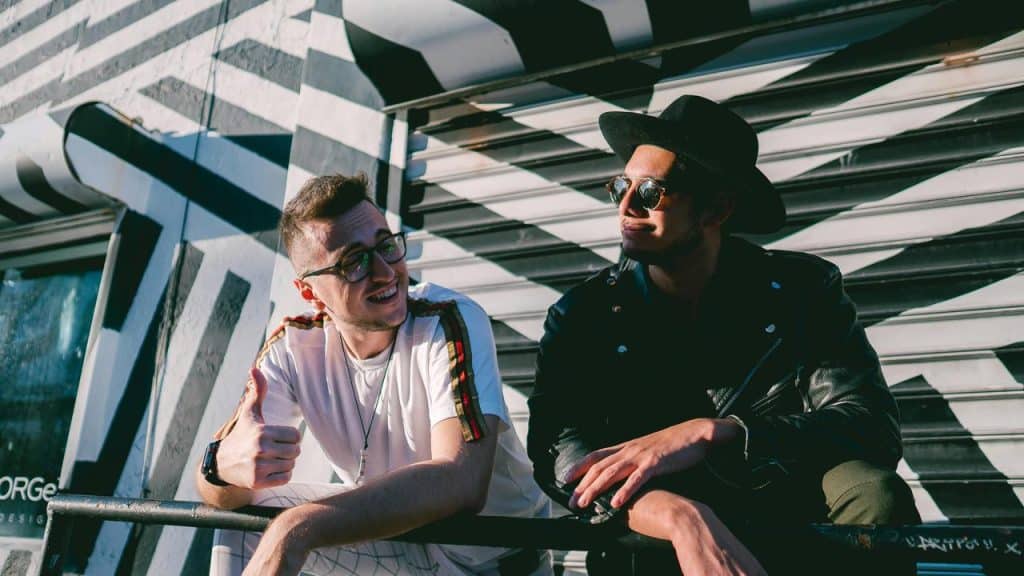 A pair of men leaning on a railing in front of a mural, one giving a thumbs-up.