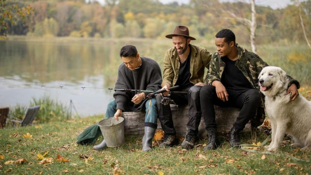 A trio of friends fishing by a lakeside with a dog.