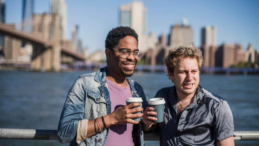 A pair of men smiling and holding coffee by the water.