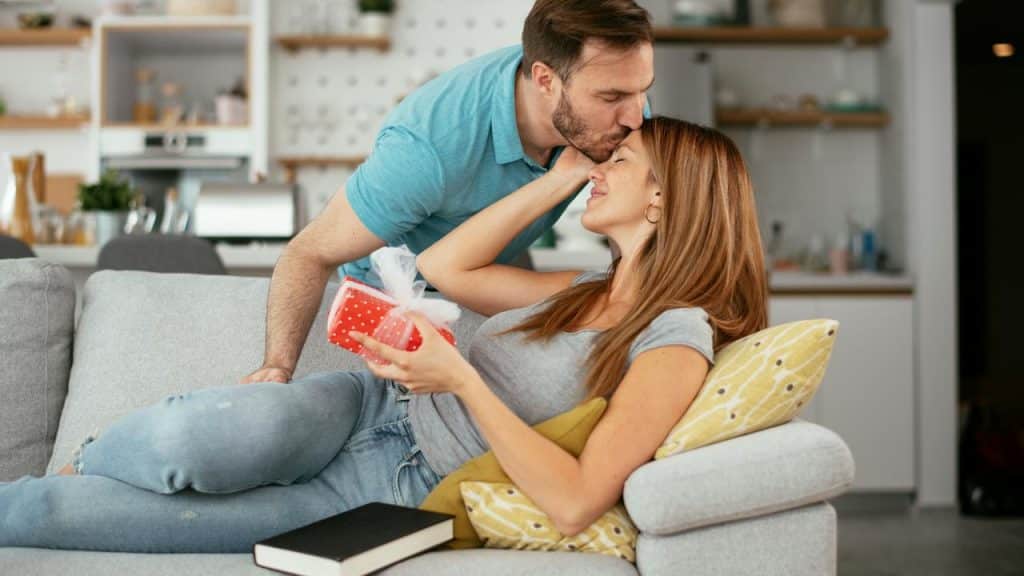 A man kissing a woman on the forehead as she holds a wrapped gift on a couch.