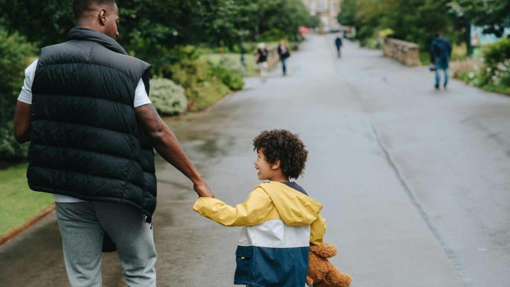A man and child walking hand in hand in a park, the child holding a teddy bear.
