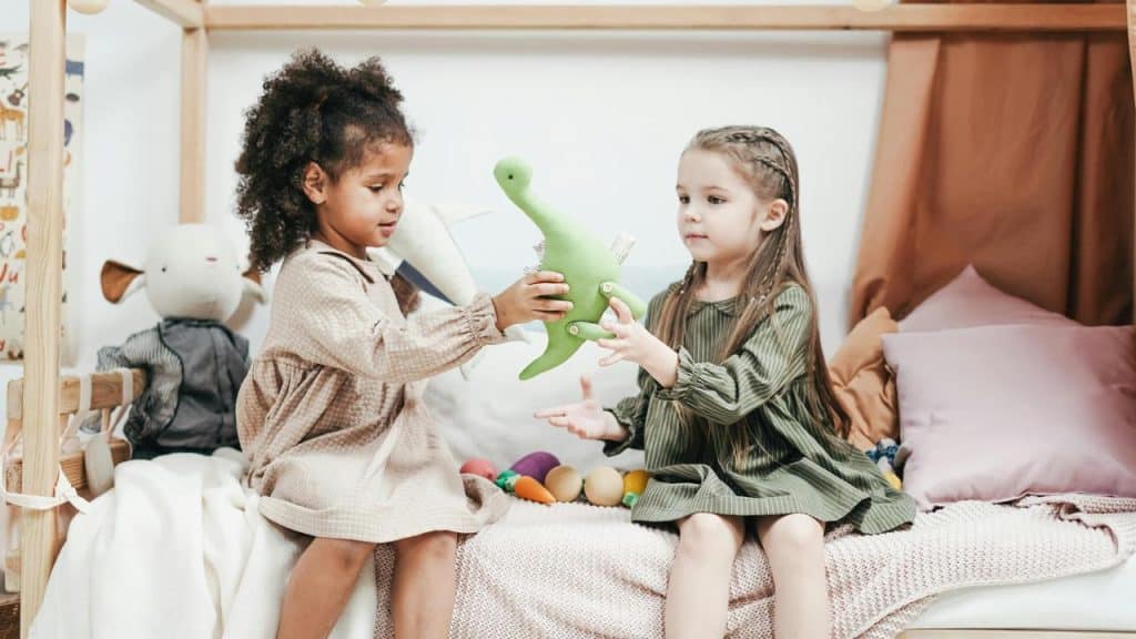 A girl handing a stuffed dinosaur to another girl on a bed.