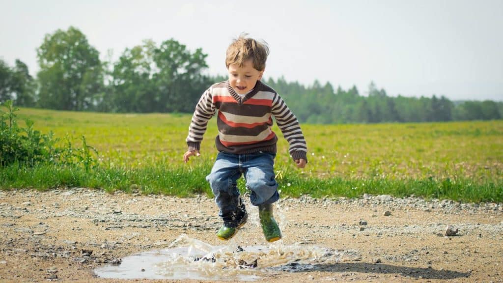 A boy jumping over a puddle on a dirt path.