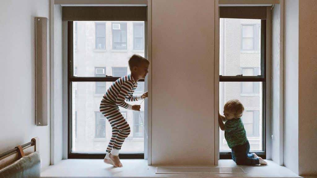 A child in striped pajamas jumping on a windowsill beside a toddler.