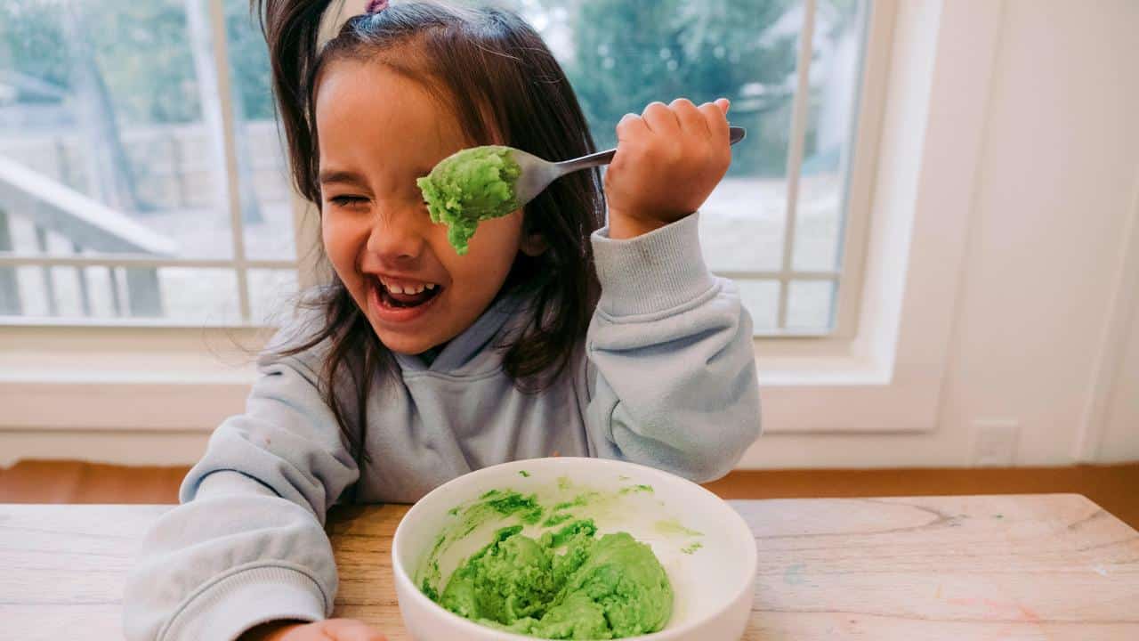 A laughing girl holding a spoon of green food.