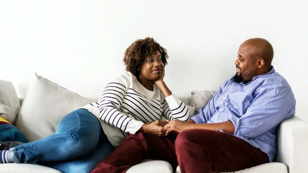 A smiling Black couple holding hands and talking on a couch.