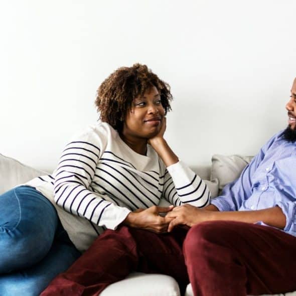 A smiling Black couple holding hands and talking on a couch.