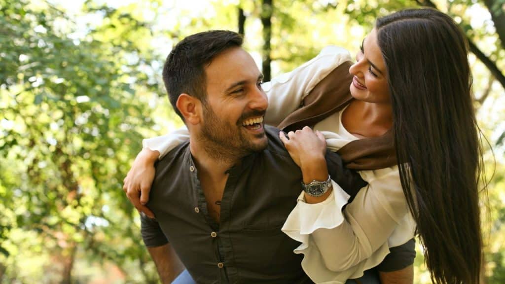 A laughing man giving a woman a piggyback ride in a sunny park.