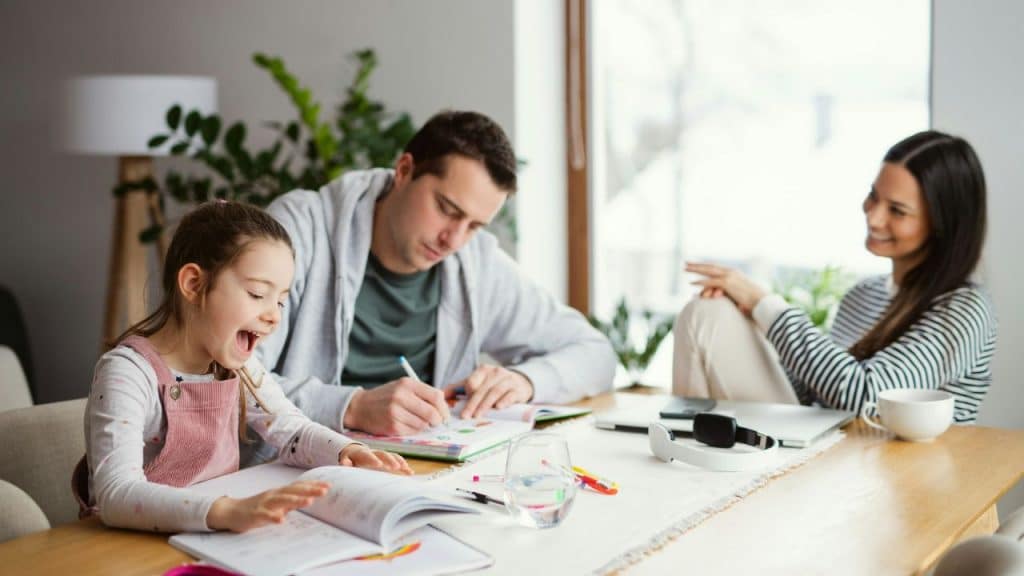 A man helps a smiling child with her homework while a woman watches.