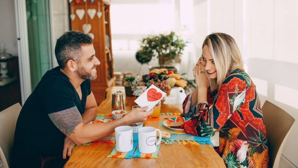 A man smiling while giving a smiling woman a card at a breakfast table.