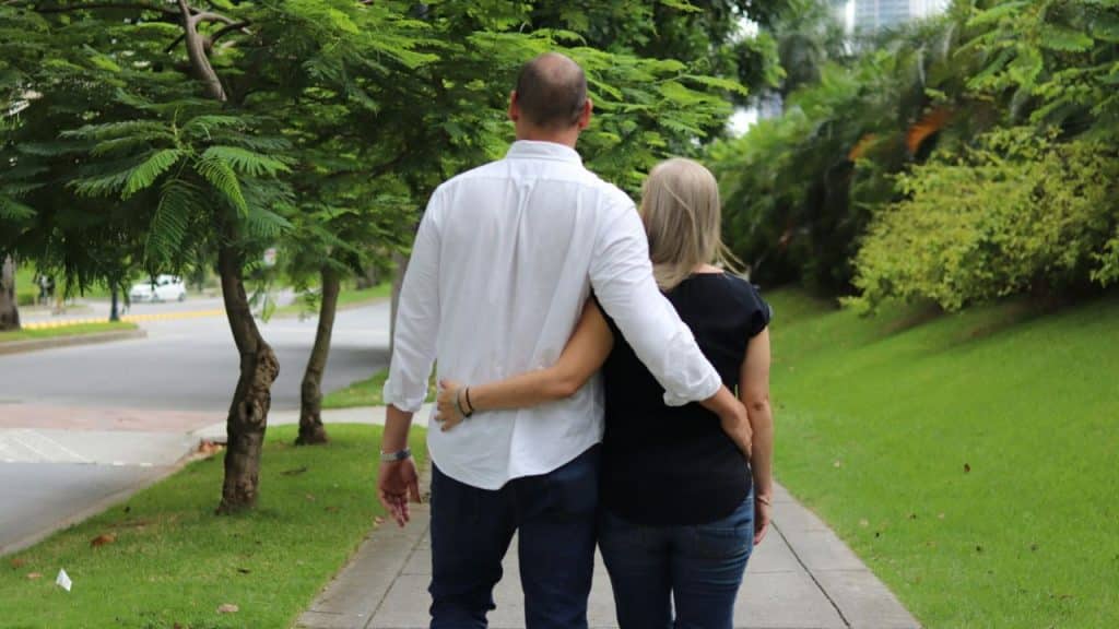 A couple from behind walking on a sidewalk with their arms around each other.