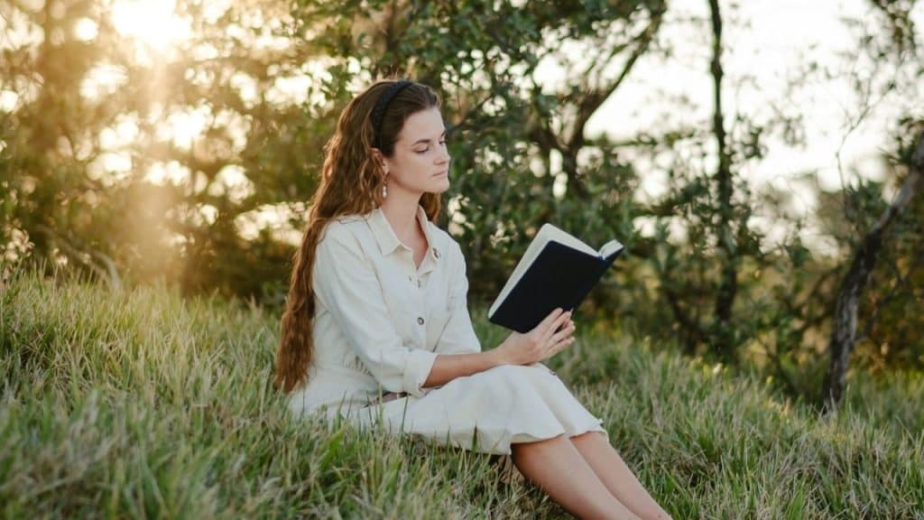 A woman in a white dress sits in a grassy field, reading a book.