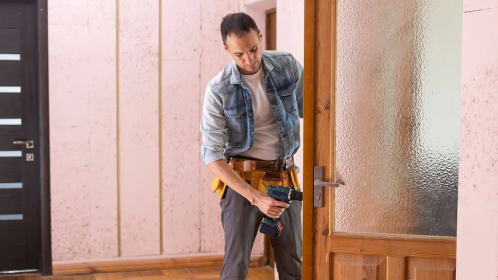 A man wearing a tool belt uses a drill to fix a door.
