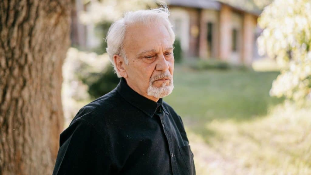 A somber, elderly man with a white beard looks off to the side.