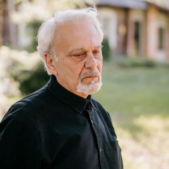 A somber, elderly man with a white beard looks off to the side.