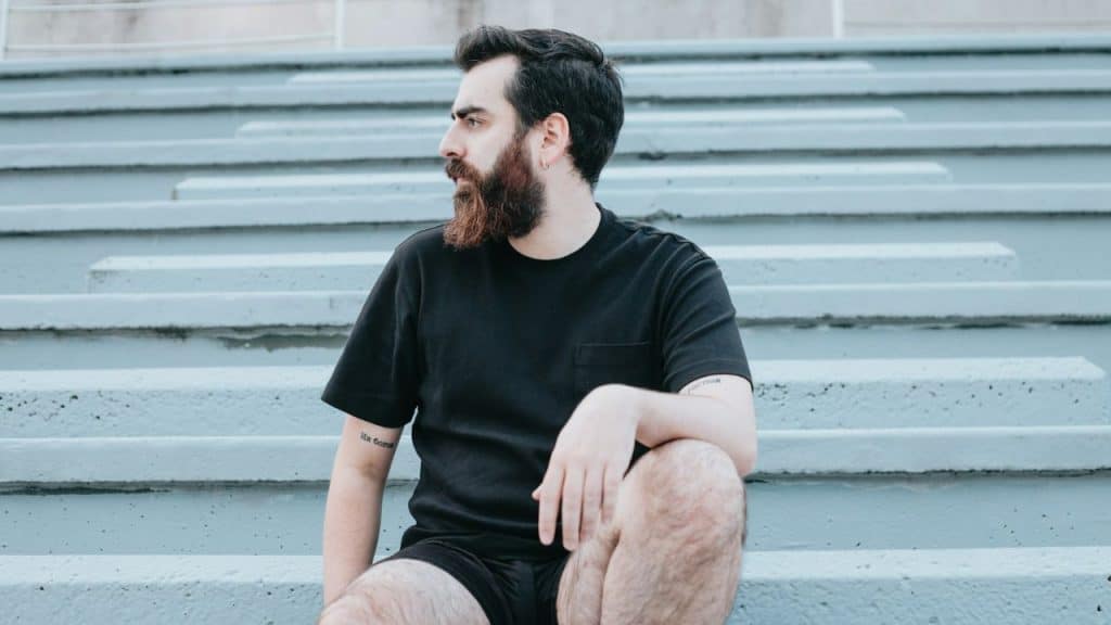 A pensive, bearded man in a black shirt sits on concrete steps.