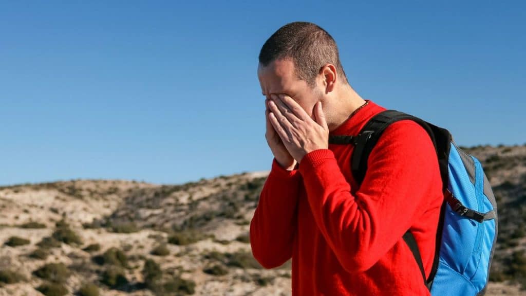 A tired man in a red sweater and backpack rubs his eyes.
