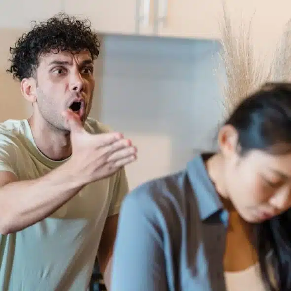 A yelling man with curly hair gestures at a woman with her head down.