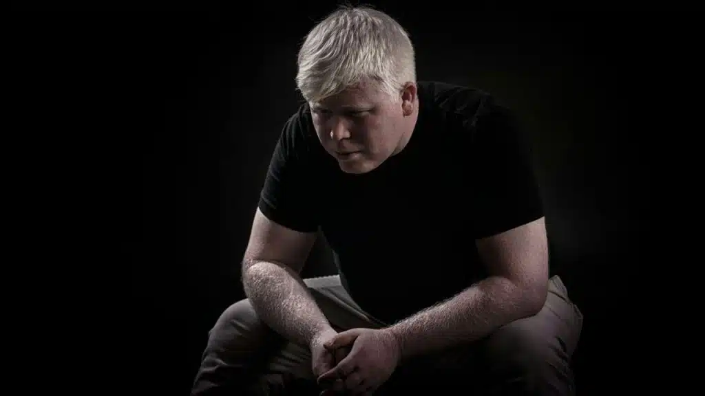 A solemn man with white hair, sitting and looking down in a dark room.