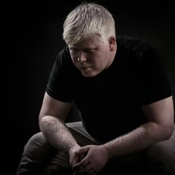 A solemn man with white hair, sitting and looking down in a dark room.