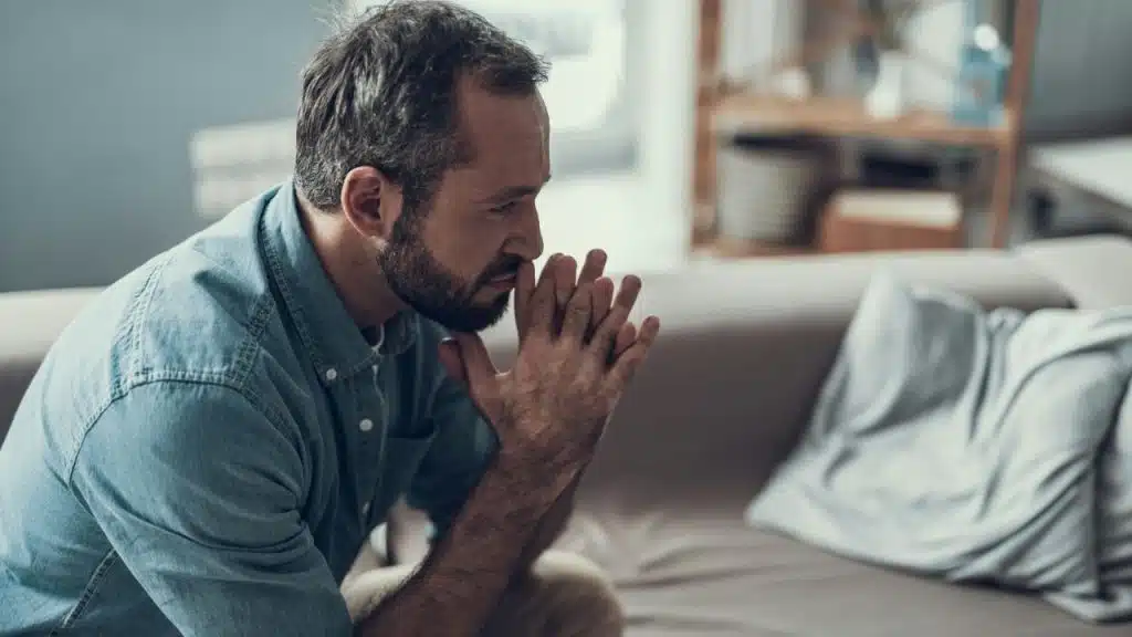 A serious man with a beard sits on a couch with his hands clasped together.