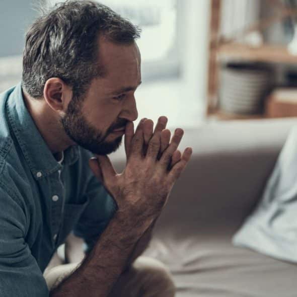A serious man with a beard sits on a couch with his hands clasped together.