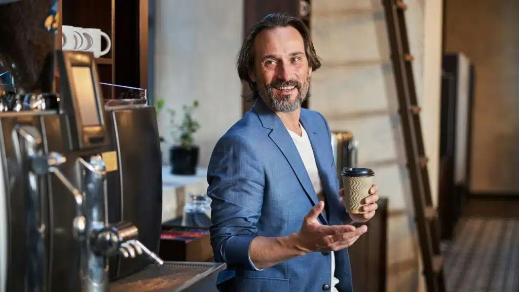 A middle-aged man with a beard, wearing a blue jacket and holding a coffee cup.