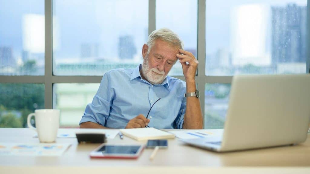 A stressed, older man with a beard sits at a desk, looking down at his notebook.