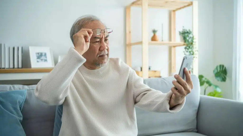 A puzzled older man with glasses and a beard looking intently at his phone.