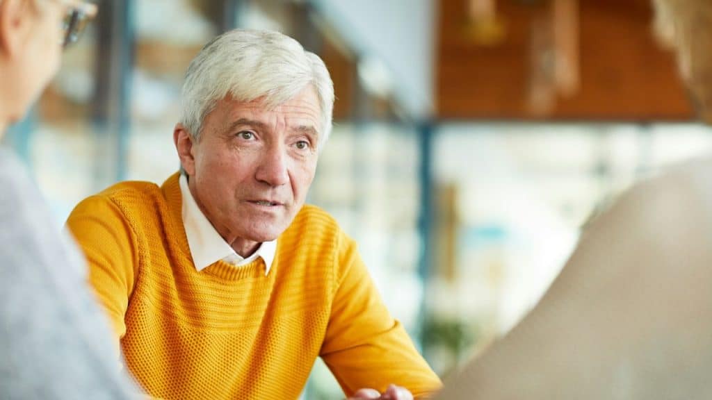 A gray-haired older man in a yellow sweater, looking concerned while speaking to someone.