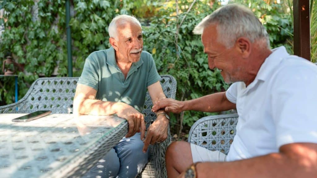 Two elderly men sitting outside at a table, one smiling at the other.