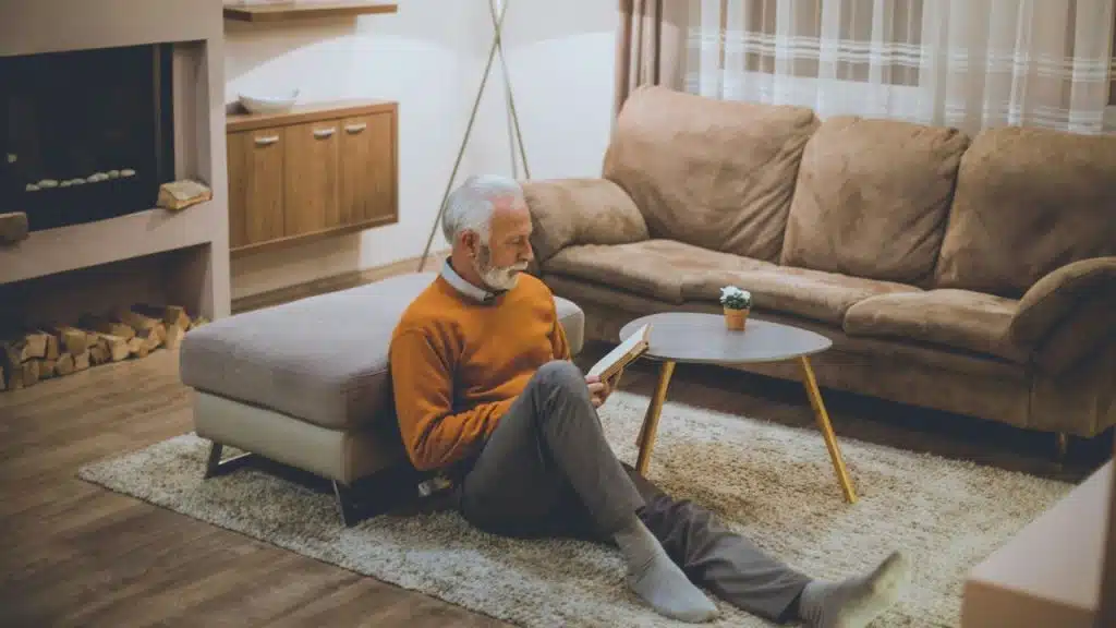 A gray-haired, bearded man in a sweater sitting on the floor, reading a book.