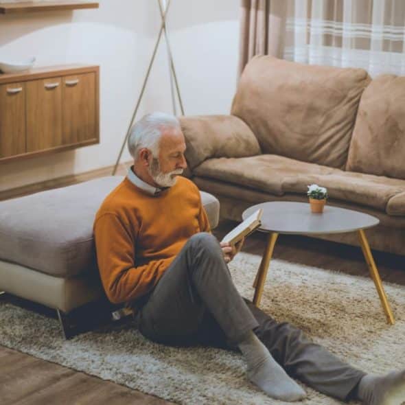 A gray-haired, bearded man in a sweater sitting on the floor, reading a book.