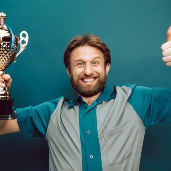 A bearded man in a two-tone shirt holds a trophy and gives a thumbs-up, smiling.