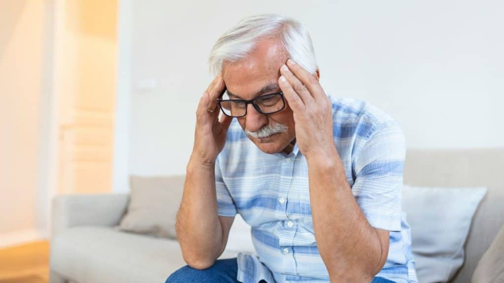 A worried elderly man with glasses and a mustache holds his head in his hands.