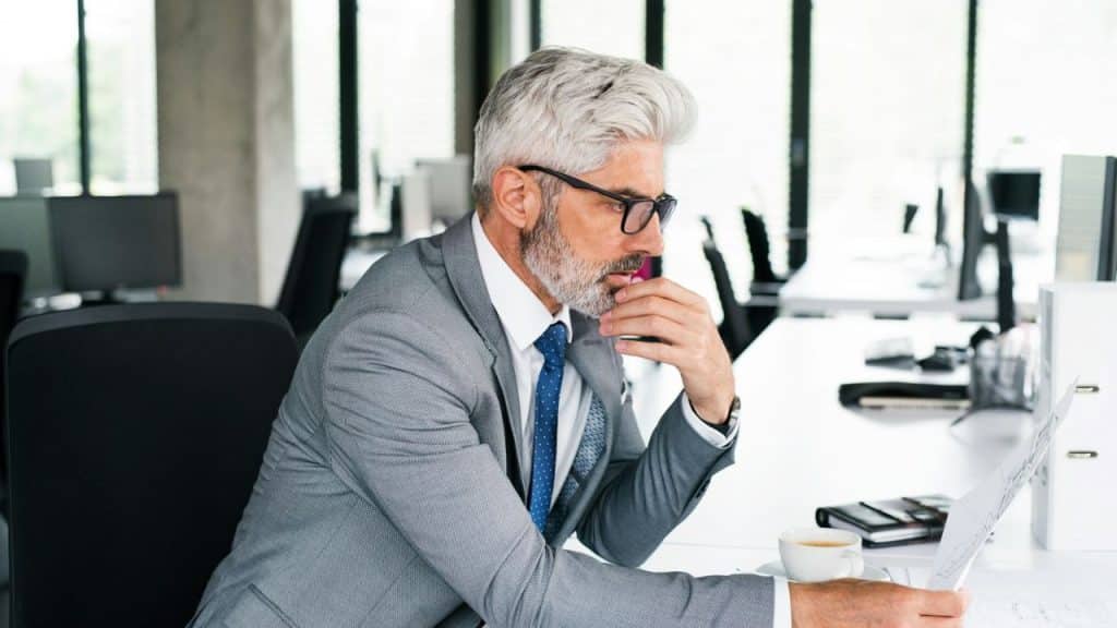 A handsome, silver-haired man in a suit reviews papers at his desk.