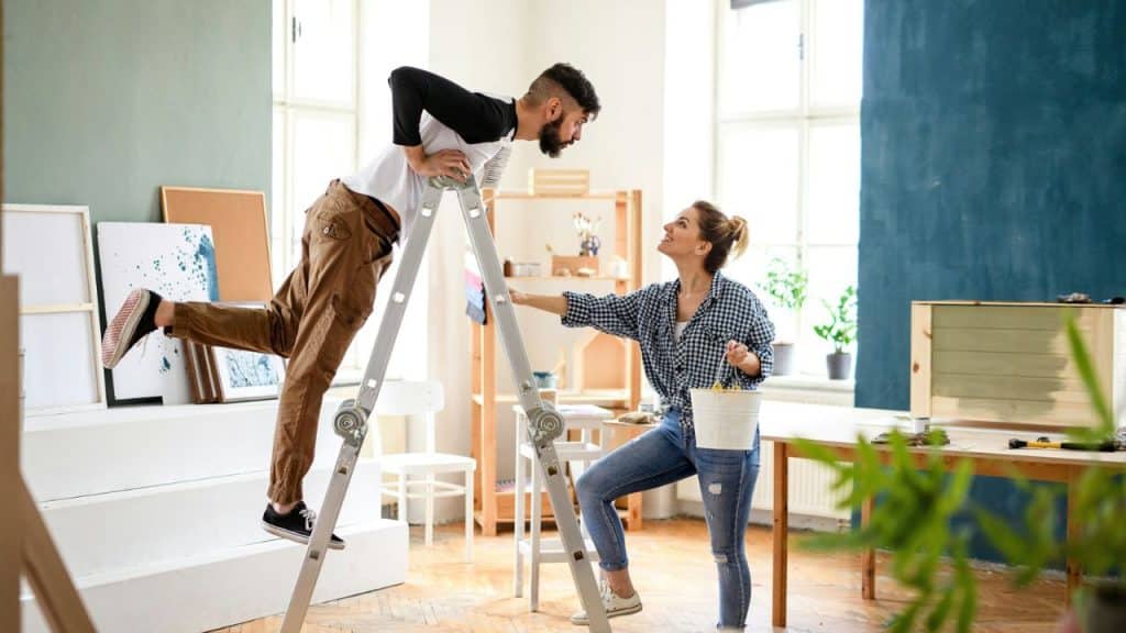 A happy couple painting a room, a man on a ladder and a woman below.