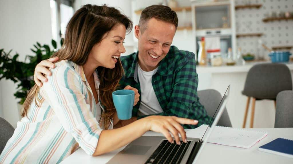 A supportive couple happily looks at a laptop together at a kitchen table.