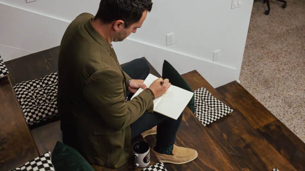 A serious man in a blazer sits on steps, writing in a notebook.