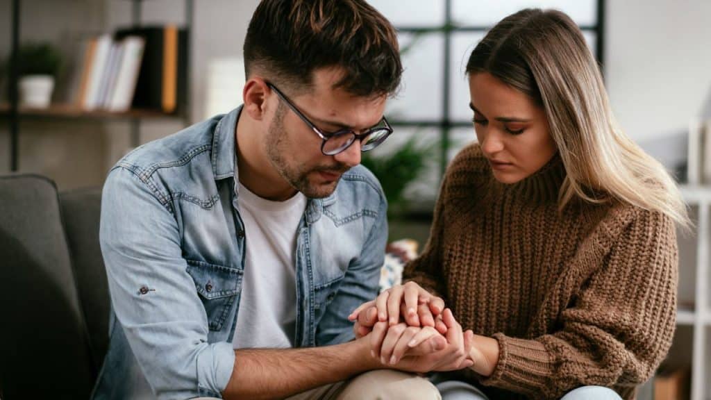 A remorseful man looks at a woman's hand, while she holds his.