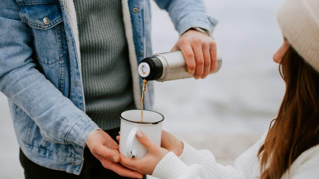 A person in a denim jacket pours coffee from a thermos into a mug for a companion.